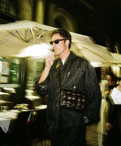 a man in black jacket and sunglasses standing under an umbrella while holding a cell phone to his ear