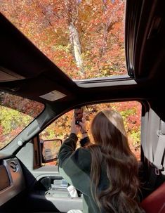 a woman taking a photo in the side mirror of a car with fall foliage behind her