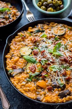 a skillet filled with lots of food on top of a black table next to bowls of olives