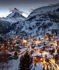 the town is surrounded by snowy mountains and lit up buildings at night in the foreground