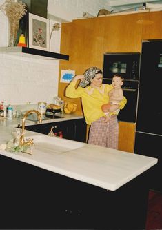 a woman holding a baby in her arms while standing next to a kitchen counter top