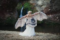 a woman in a white dress is standing near a waterfall with her wings spread out