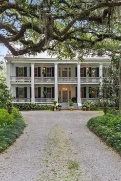 a large white house surrounded by trees and bushes in the middle of a gravel road