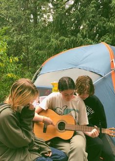 three young people sitting in front of a tent playing guitar