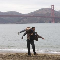 a man carrying a child on his back while walking along the beach in front of the golden gate bridge