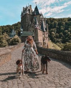 a woman is walking with two dogs in front of an old castle on a cobblestone road