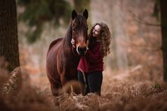 a woman standing next to a brown horse in a forest with tall grass and trees