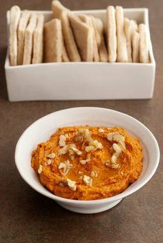 a white bowl filled with hummus next to a container of pita bread sticks