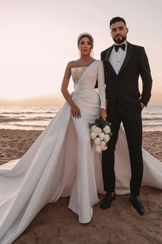 a man and woman standing on top of a beach next to each other wearing tuxedos