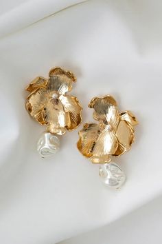 two gold flower shaped earrings sitting on top of a white cloth covered table next to a pair of pearls