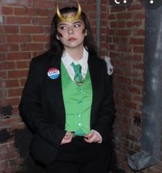 a woman in a green shirt and black suit with horns on her head standing next to a brick wall