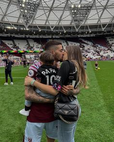 two people kissing each other in front of a soccer field with fans watching from the stands