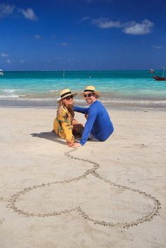 two people sitting on the beach with a heart drawn in the sand