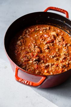 a red pot filled with chili and meat on top of a white countertop next to a wooden spoon