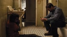 a man sitting in a bathroom next to a woman on the floor looking at her cell phone