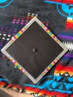 a black square on a colorful blanket with a button in the center and an embroidered border around it