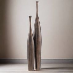 two tall metal vases sitting next to each other on a wooden floor in front of a wall