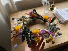 a hand is holding a wreath made out of dried flowers on a table next to an open book