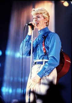 a man in blue shirt and white pants holding a microphone up to his ear while standing on stage