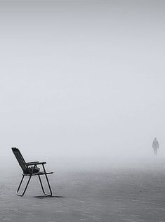 an empty chair in the middle of a foggy field with a person standing behind it