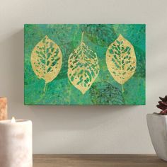 three green and gold leaf paintings on a wall above a wooden table next to a potted plant