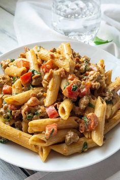 a white plate topped with pasta covered in sausage and tomato sauce next to a glass of water