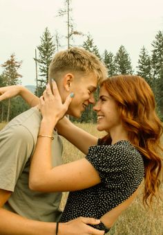 a young man and woman embracing each other in the middle of an open field with pine trees behind them