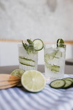 two glasses filled with cucumber and ice on top of a table next to sliced limes