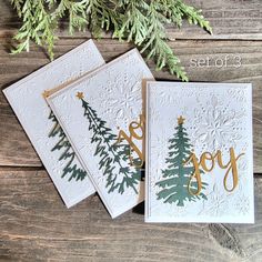 three cards with christmas trees on them and the words joy written in gold foil are sitting next to evergreen branches