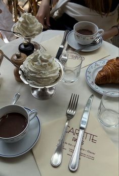 a table topped with two cups of coffee and desserts next to silver utensils