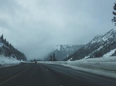 an empty road with snow on the mountains in the back ground and trees along side