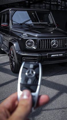 a hand holding a remote control in front of a black mercedes g - class car