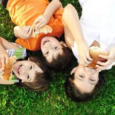 three children laying on the grass eating hotdogs and hamburgers with their hands