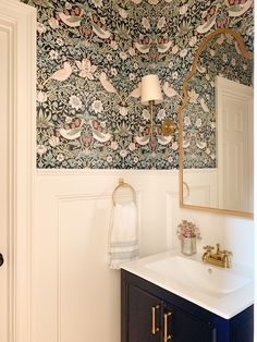 a bathroom with floral wallpaper and gold faucet, sink, mirror and towel rack