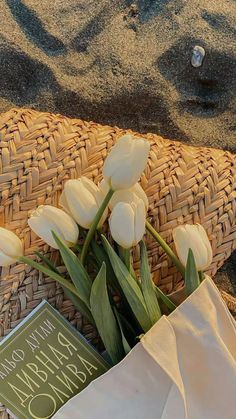 white tulips are in a basket on the sand with a book about happiness