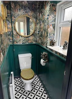 a bathroom with green walls and black and white tile flooring, along with a gold mirror above the toilet