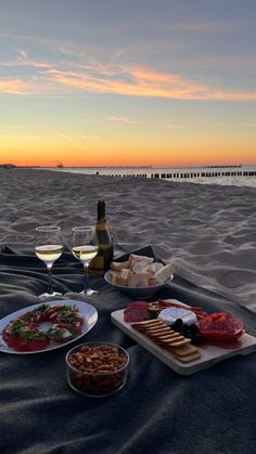 a bottle of wine and two plates of food on a blanket at the beach with sunset in the background