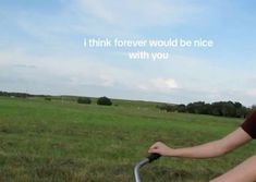 a woman riding on the back of a bike down a lush green field next to a forest