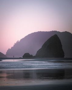 a person is standing on the beach near some rocks and water at sunset or dawn