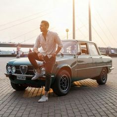 a man sitting on the hood of an old car in front of a bridge at sunset