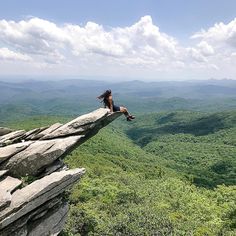 a woman sitting on top of a large rock next to a lush green forest covered hillside