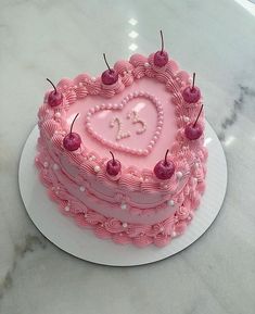 a pink heart shaped cake with cherries on top sits on a white plate in front of a marble countertop