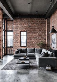 An industrial-style living room with exposed brick walls painted in a muted grey tone. The room features a grey sectional sofa, black metal accents, and modern light fixtures. Concrete flooring and large windows with a city view add to the urban vibe. Living Room With Exposed Brick, Industrial Living Rooms, Industrial Living Room Ideas, Industrial Living Room Design, Latest Decorating Trends, Industrial Living Room, Gray Living Room Design, Contemporary Loft, Grey Sectional Sofa