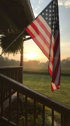 an american flag on the porch of a country house at sunset or sunrise with grass and trees in the background