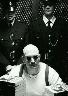 two men in uniform are sitting at a table with stacks of books and one man is wearing sunglasses