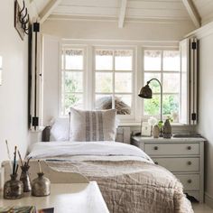 a bed sitting under two windows next to a desk with a lamp on top of it