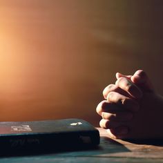 two hands clasped over a book sitting on a wooden table in front of the sun