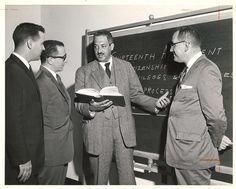 three men standing in front of a blackboard while one man holds an open book