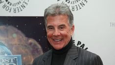 a smiling man in a suit and turtle neck sweater standing next to a post office sign