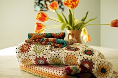 three crocheted blankets stacked on top of each other next to a vase with tulips